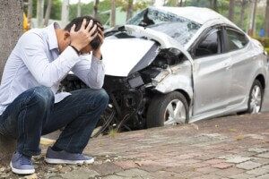 man crouched down with his hands on his head in distress, a crushed car behind him